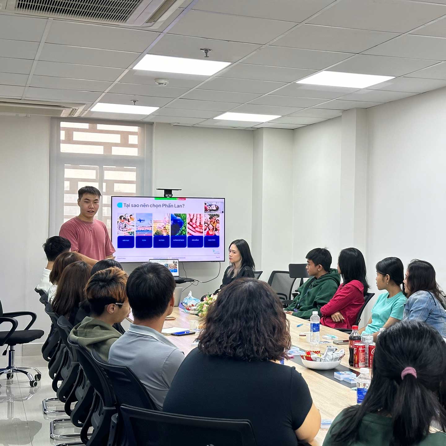 A man is standing and presenting to a group of people seated around a conference table. A screen displays a presentation with colorful graphics. The room has a bright, modern interior with a large window in the background.