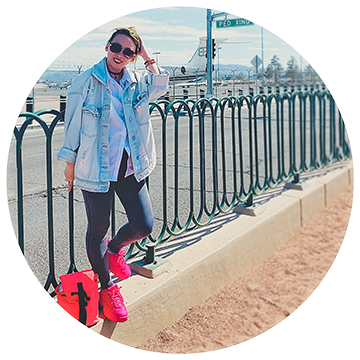 Person wearing bright pink sneakers and a denim jacket stands on a sidewalk, leaning against a metal fence. A red bag is on the ground nearby. A street sign and road are visible in the background, with a clear sky above.