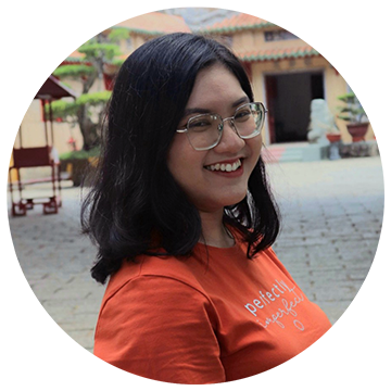 A woman with long black hair and glasses smiles while standing outdoors. She is wearing an orange T-shirt with white text. The background features a courtyard with trees and traditional architecture.