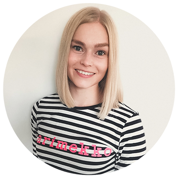 A person with straight blonde hair smiling, wearing a black and white striped shirt with pink text reading 