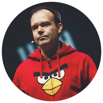 A person wearing a red hoodie with an Angry Birds character design is speaking on stage. They have a microphone headset and appear to be giving a presentation. The background is dark and blurred.