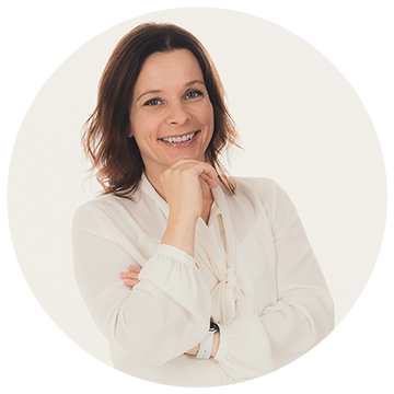 Smiling woman with shoulder-length brown hair, wearing a white blouse, poses with her hand under her chin against a light background.