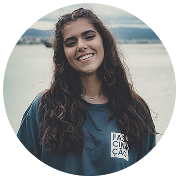 A young woman with long curly hair smiles at the camera, standing outdoors. She wears a blue shirt with the word "FASCINAÇÃO" printed on it. The background is blurred, with natural scenery.