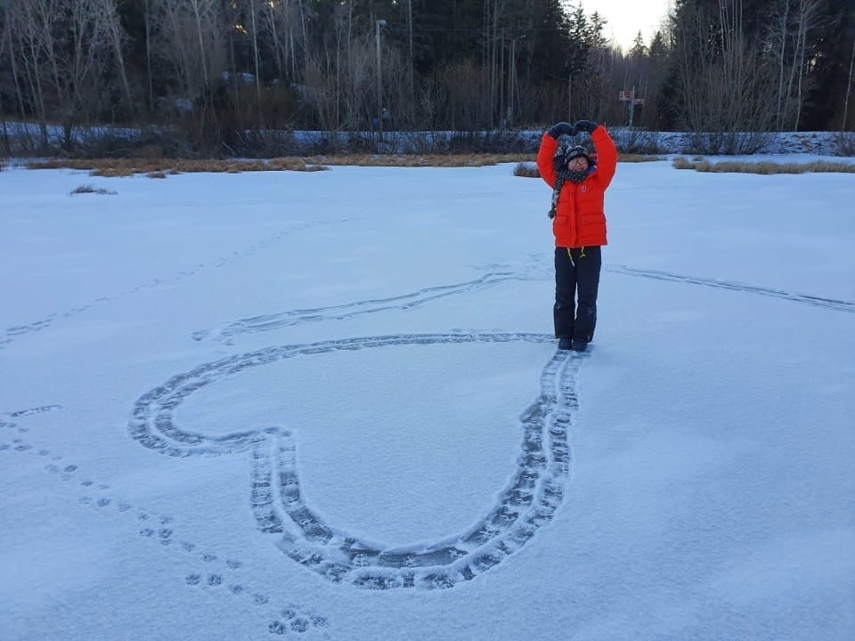 A child in a red coat stands on a snowy field, holding their hands above their head. A large heart shape is drawn in the snow around them, with footprints visible. Trees line the background under a clear sky.