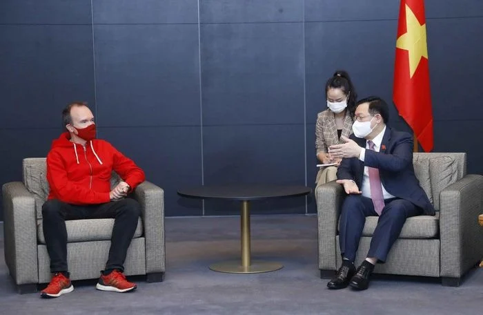 Two people sit facing each other in armchairs, both wearing face masks. The person on the left wears a red hoodie, while the person on the right is in a suit gesturing with one hand. A Vietnamese flag is visible in the background.
