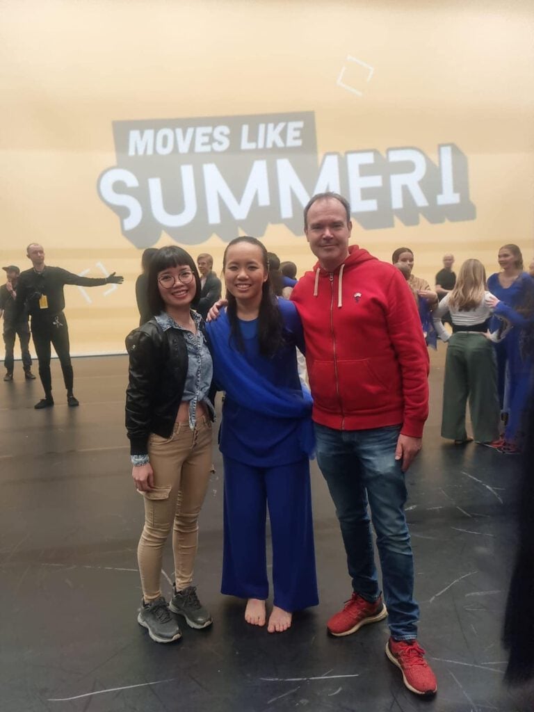 Three people pose together onstage. The background features a large sign reading "MOVES LIKE SUMMER!" Other individuals are visible in the background. The central person wears a blue outfit, flanked by one in a black jacket and another in a red hoodie.