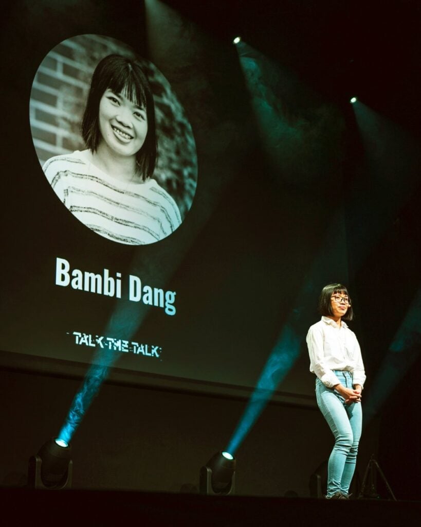 A woman stands on stage under a spotlight, smiling. On the screen behind her is a projected photo with the name "Bambi Dang" and the text "Talk the Talk." The background is dark with additional spotlights.