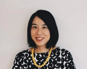 A person with shoulder-length black hair smiles at the camera. They are wearing a patterned black and white top and a yellow beaded necklace, standing against a plain background.