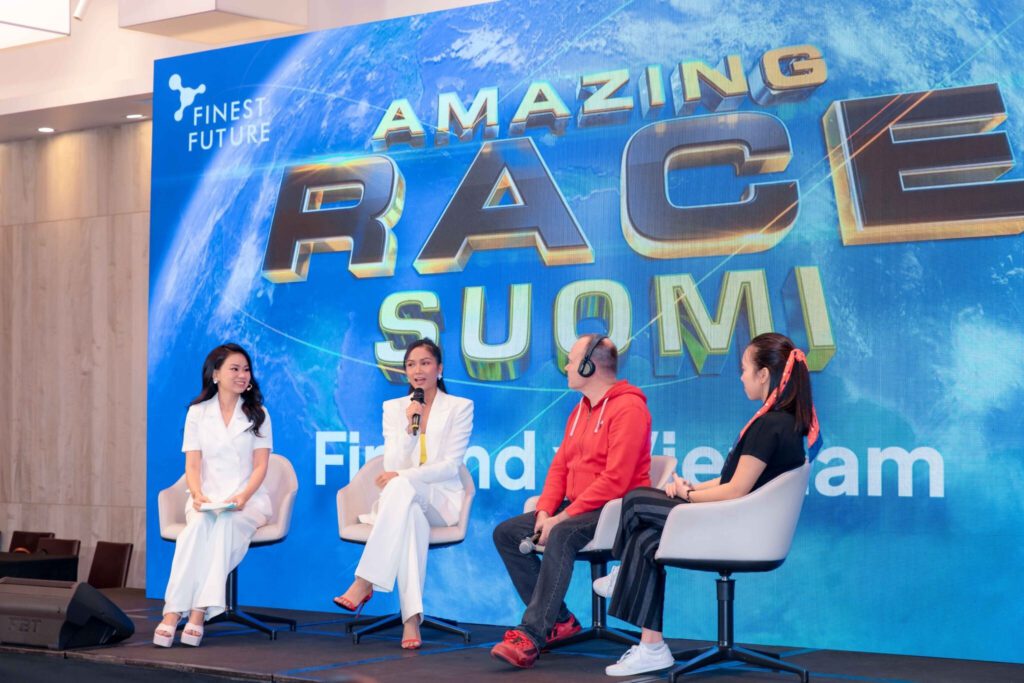Four people are seated on a stage, engaged in conversation. Behind them is a large sign that reads "Amazing Race Suomi." They appear to be part of a panel or discussion event.
