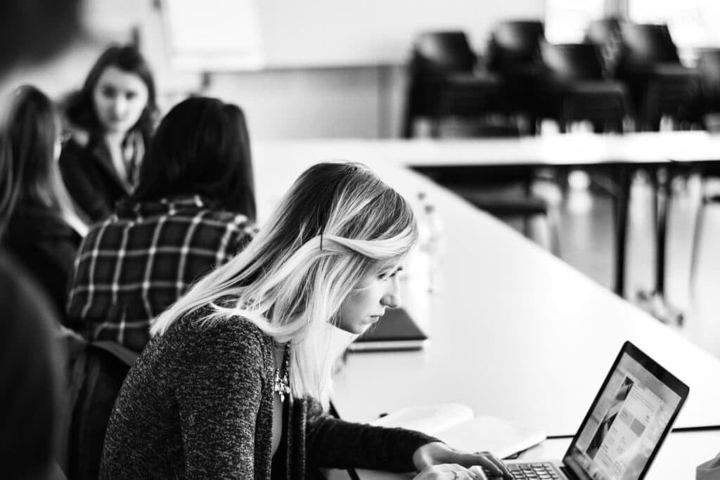 A black and white photo of a woman with blonde hair working on a laptop at a long table. In the background, three other people are engaged in conversation. The room is spacious with empty chairs visible.