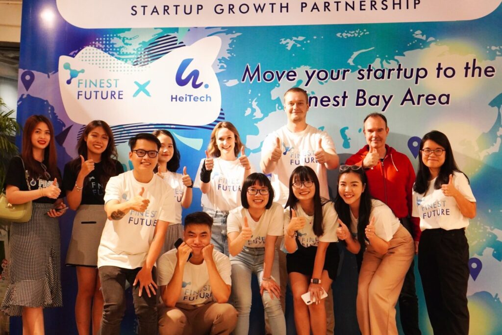 A diverse group of people wearing "Finest Future" T-shirts poses with smiles and thumbs up in front of a banner reading "Startup Growth Partnership" and "Move your startup to the Finest Bay Area.