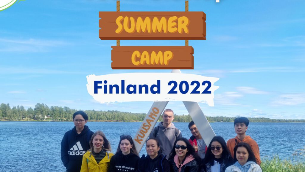 A group of people stand by a lake under a clear blue sky, with a sign above reading "Summer Camp, Finland 2022." Trees are visible in the background.