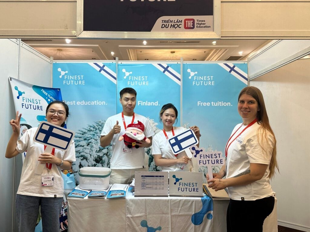 Four people stand in front of a booth with "Finest Future" branding. Two hold Finnish flags, and one holds a plush toy. They're smiling and wearing white shirts with red lanyards. A snowy background is partially visible.