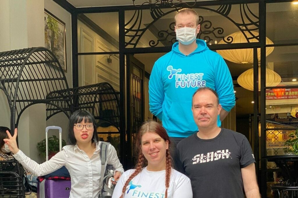 Group of four people posing in front of a building entrance. One person wears a blue hoodie, another a white shirt, another a black shirt, and one is holding up a peace sign. A pink suitcase is visible in the background.