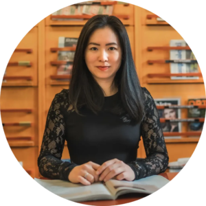 A woman with long black hair sits at a table with a book open in front of her. She is wearing a black lace top. The background features a wooden shelf with books and magazines.