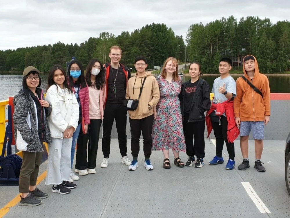 A group of ten people standing on a dock with a picturesque lake and forest in the background. They are smiling and dressed in casual clothing. The scene appears relaxed and friendly, with overcast skies.