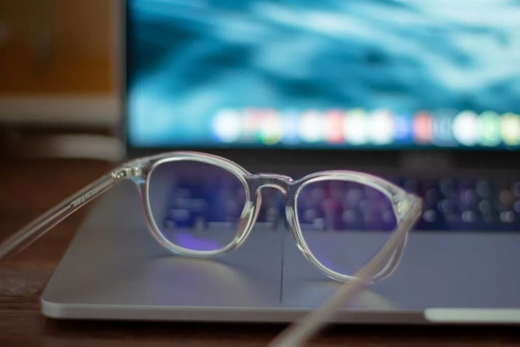 A pair of transparent glasses rests on a partially open laptop. The laptop screen displays a blurred, wave-like blue and green background with a row of colorful icons at the bottom. The scene appears focused on minimalism and technology.