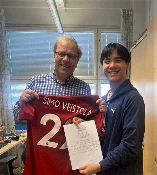 Two people smiling indoors. One holds a red sports jersey with the name "Simo Veistola" and the number 22. The other person holds a piece of paper. They are standing in a room with a desk and windows in the background.