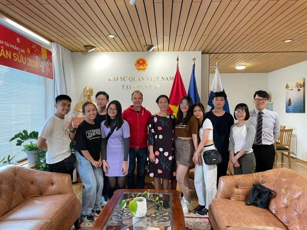 A group of eleven people stands smiling in an office with leather couches and flags in the background. The office has wooden ceilings and a plaque on the wall. They are dressed casually and appear to be posing for a group photo.