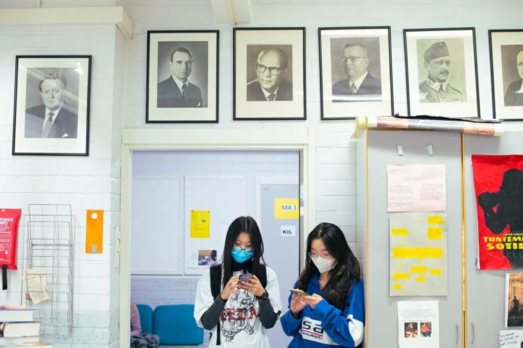 Two people wearing masks stand in a room with framed portraits on the wall above them. They are looking at their phones, and there's a mix of books, papers, and posters around them. The environment suggests a classroom or study area.