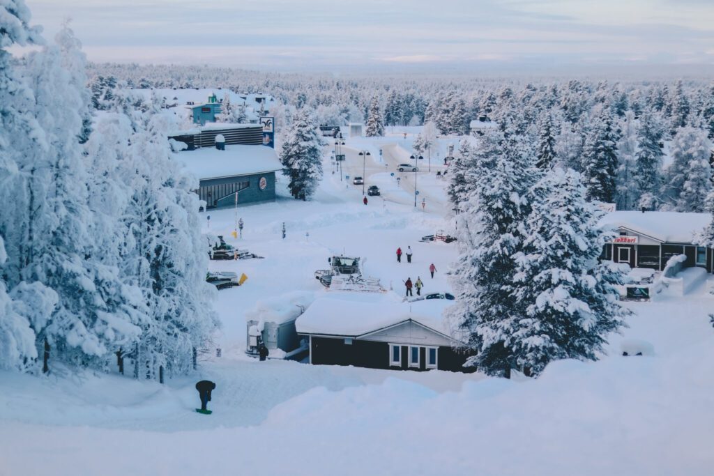 A snowy village scene with snow-covered trees and buildings. People are walking on snow pathways between structures. The landscape is blanketed in snow under a cloudy sky, creating a serene winter atmosphere.