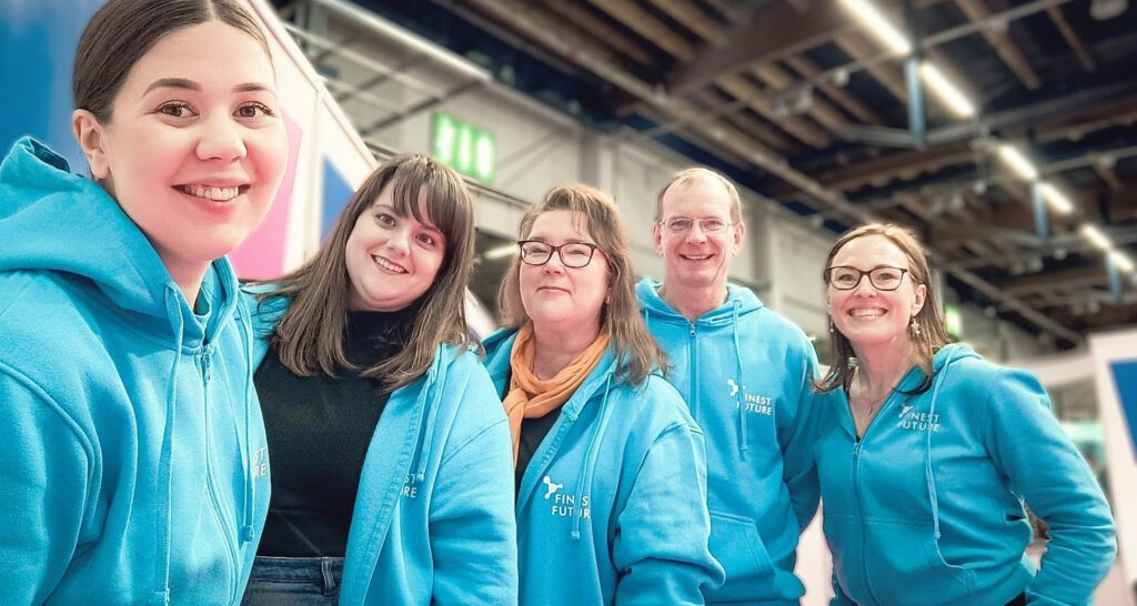 A group of five people wearing matching blue hoodies, some with glasses, are smiling indoors. They stand closely together under a ceiling with exposed beams.
