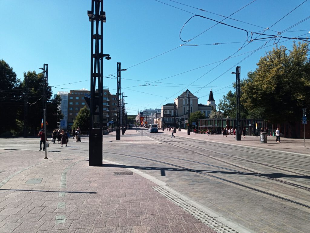 A sunny day scene showcasing a spacious street with tram tracks, a few pedestrians walking, and buildings in the background. Tall trees line the sides, and a clear blue sky is visible above.