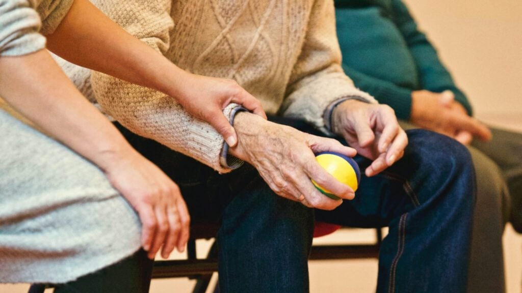 A person in a sweater sits holding a colorful ball, while another person's hand rests gently on their arm. A third person is seated nearby, partially visible. The scene suggests a supportive or caregiving environment.