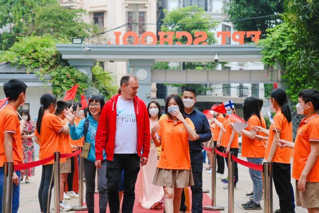 A group of students in orange shirts welcomes two people walking down a path, lined with students waving flags. The entrance to a school with the letters partially visible is in the background.
