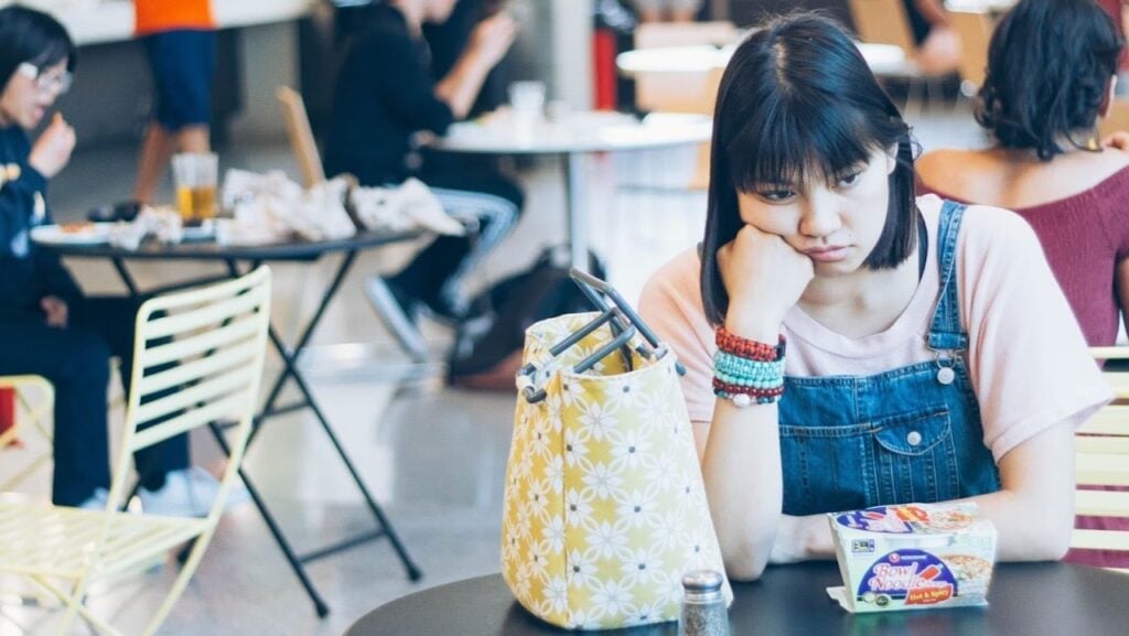 A young woman in denim overalls sits at a cafe table, resting her chin on her hand, looking thoughtful or bored. A floral bag and a yogurt cup are in front of her. Other people are seated in the background.