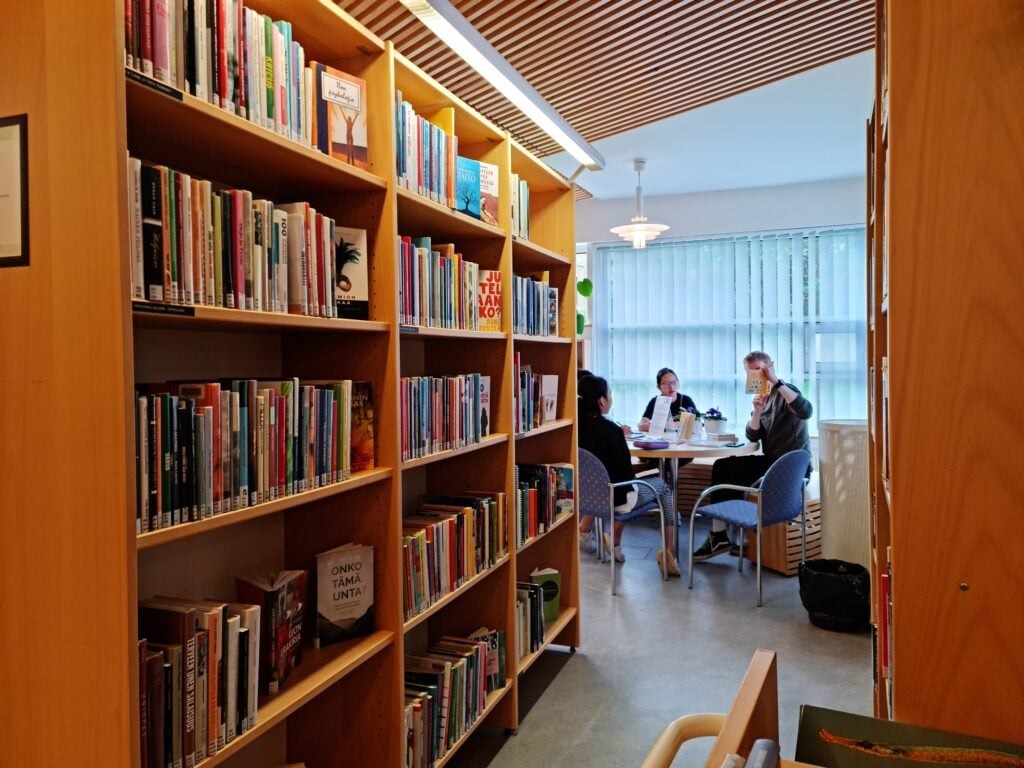 Celebrating Helsinki's Oodi Library - A Modern Marvel for Students and Culture Enthusiasts 3 A cozy library with wooden shelves filled with books on the left. In the background, four people are seated at a round table, engaged in conversation. Natural light streams in from large windows, creating a warm atmosphere.