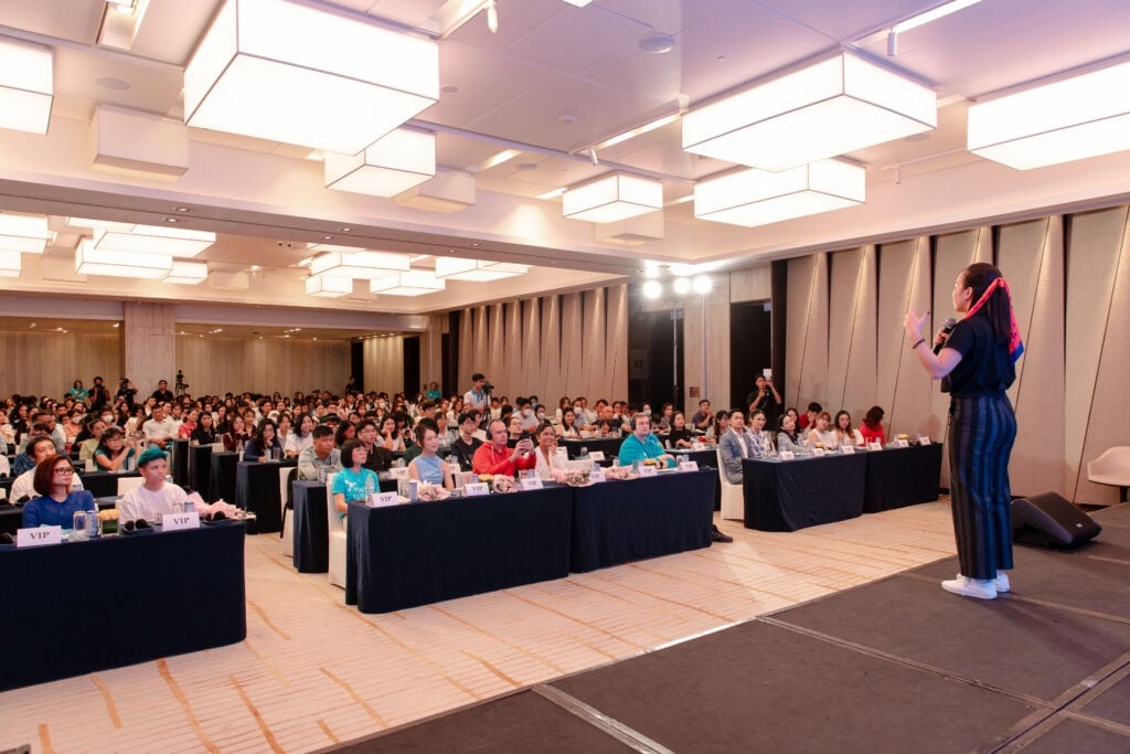 Cuộc đua kỳ thú Phần Lan x Việt Nam: Hợp nhất giáo dục và phiêu lưu 2 A speaker in a striped outfit addresses a large audience in a conference hall. Attendees are seated at tables filled with notepads and drinks. The room features modern lighting fixtures and a neutral color scheme.