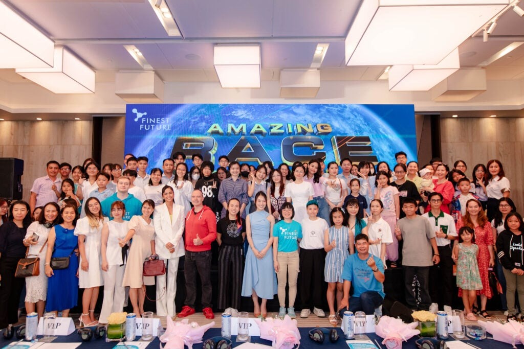 Cuộc đua kỳ thú Phần Lan x Việt Nam: Hợp nhất giáo dục và phiêu lưu 4 A large group of people stands together in front of a stage backdrop that reads "Amazing Race." They are smiling and dressed in a variety of casual and semi-formal clothing. The room features modern lighting and decor.