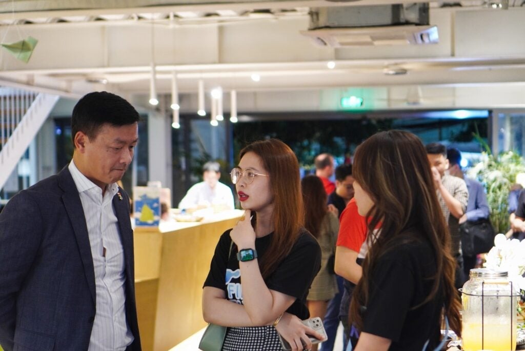 Finest Future’s Entrepreneurship Partnership Event an Overwhelming Success 4 A group of people are gathered in a modern, well-lit indoor space. A man in a suit listens to two women, one wearing glasses and both in casual black tops. Other people are in the background, engaged in conversation.