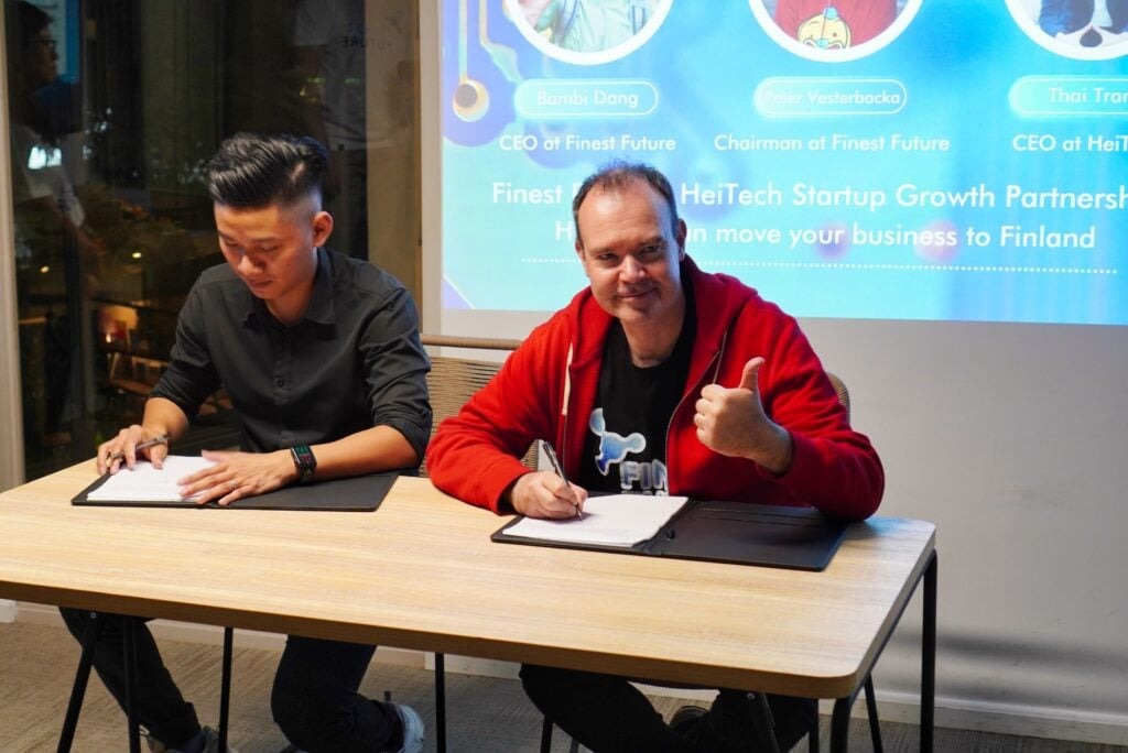 Finest Future’s Entrepreneurship Partnership Event an Overwhelming Success 2 Two men sit at a table signing documents. The man on the right, wearing a red jacket, gives a thumbs-up and smiles. Behind them is a screen displaying company and sponsor details.