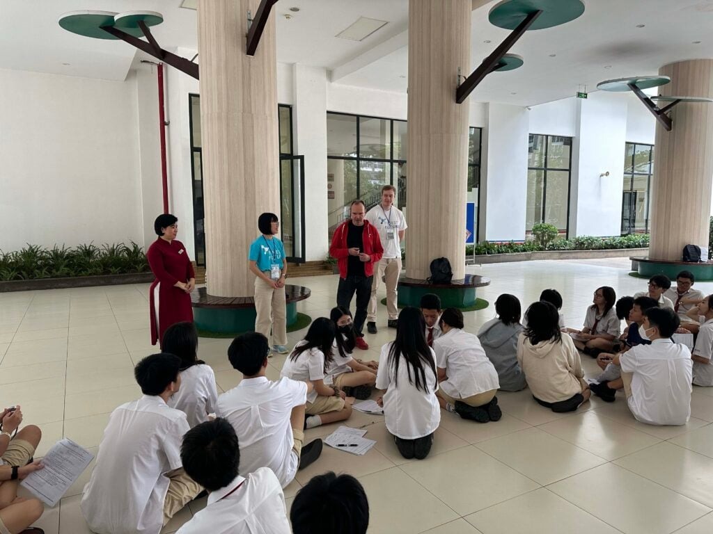 Đưa giáo dục Phần Lan đến Việt Nam 7 A group of students sit on the floor in a spacious, bright room with tall columns. Three people stand in front, with one speaking to the seated group. Papers are scattered on the floor, and there's greenery in the background.