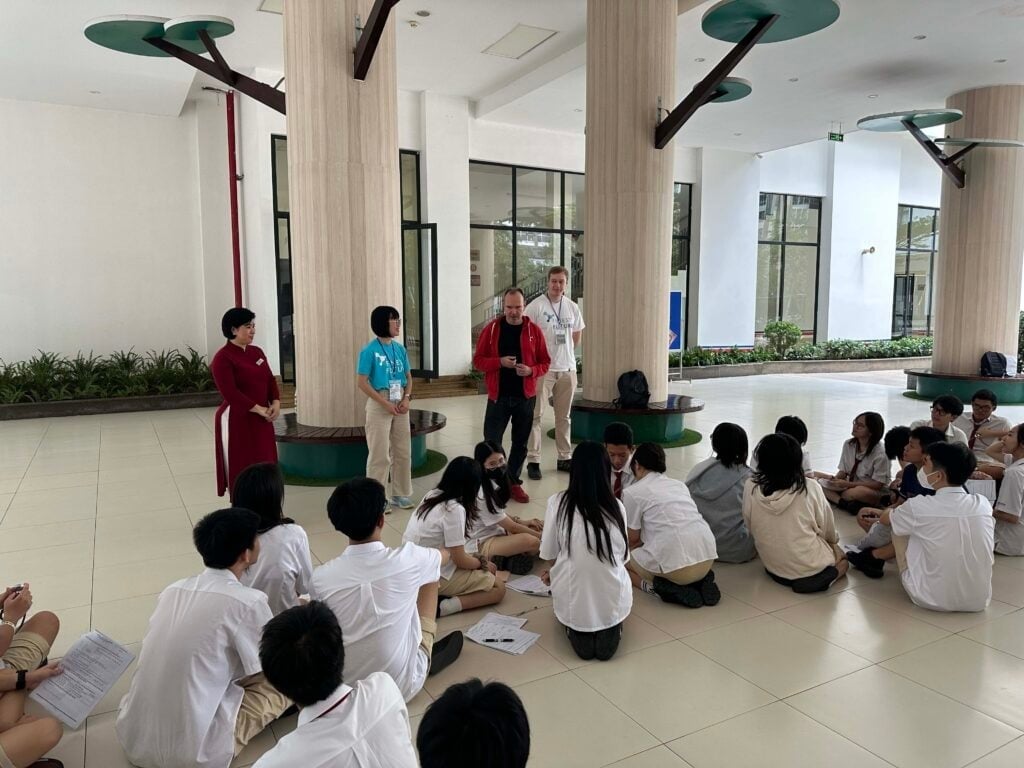 Bringing Finnish Education to Vietnam 7 A group of students in white uniforms sit on the floor in a spacious, well-lit area with columns and tree-like structures. Two adults, one in a red jacket, stand with two students at the front, engaging the group in discussion.