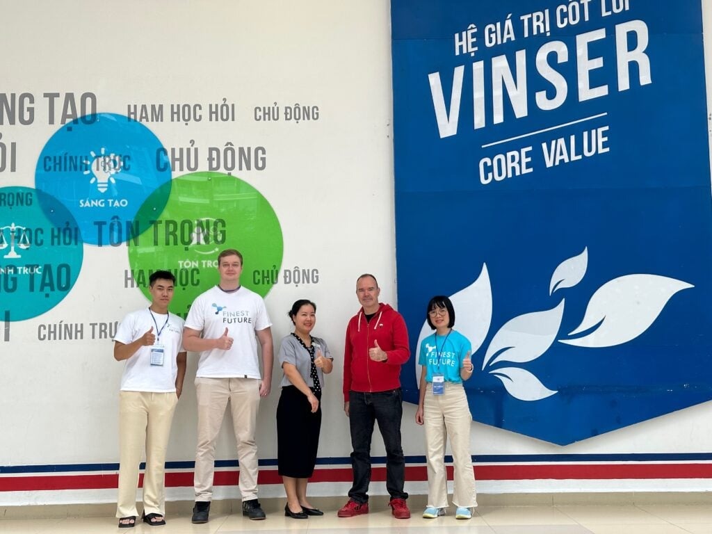 Đưa giáo dục Phần Lan đến Việt Nam 6 Five people are standing in front of a wall displaying the text "Hệ Giá Trị Cốt Lõi VINSER Core Value." Some members are wearing matching t-shirts. They are all smiling and showing thumbs up. The wall has green and blue graphic elements.