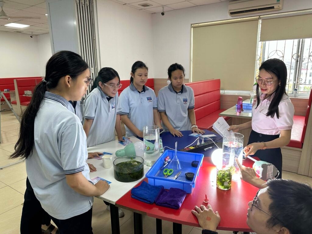 Bringing Finnish Education to Vietnam 5 A group of students and a teacher are gathered around a table in a classroom, engaged in a hands-on science activity involving containers of water and plants. The teacher is holding papers while students observe and discuss.