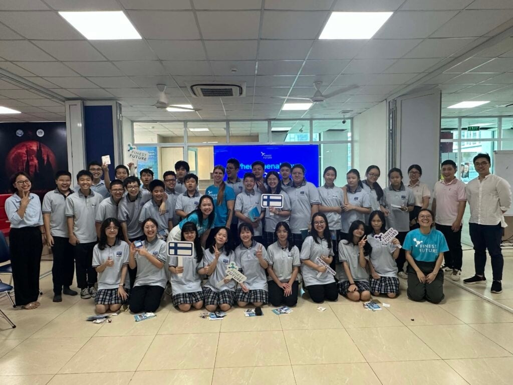 Đưa giáo dục Phần Lan đến Việt Nam 4 A large group of students and adults in a classroom, all wearing matching gray polo shirts, except for three individuals in different attire. They're smiling and holding various signs in front of a screen displaying "the arena.