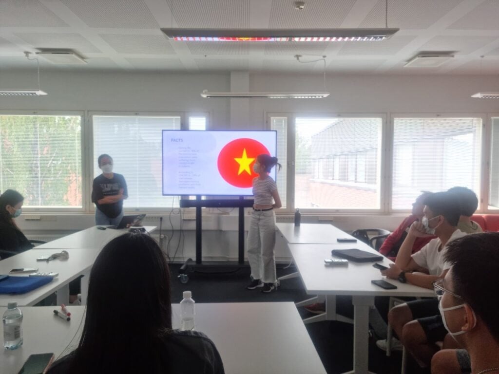 HS2021 Diary: The first Summer Camp ever in Finland! 3 A group of people sitting at tables in a bright room. Two individuals stand near a screen displaying the Vietnamese flag and a list titled "FACTS". Some participants are wearing face masks, and large windows provide natural light.