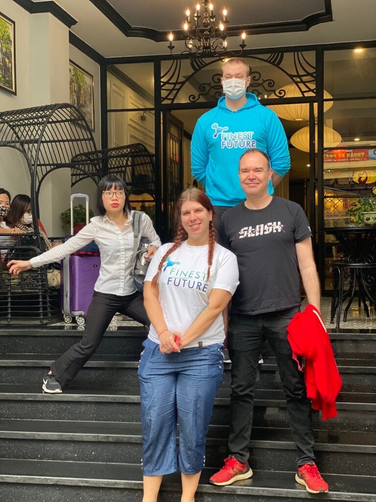 Finest Future Set to Acquire Finnish Language Education Company DreamEd Oy 3 Four people are standing together on the steps of a building entrance. One woman is seated on a railing with a purple suitcase beside her. The man on the top step is wearing a mask and a bright blue hoodie. The group appears cheerful and relaxed.