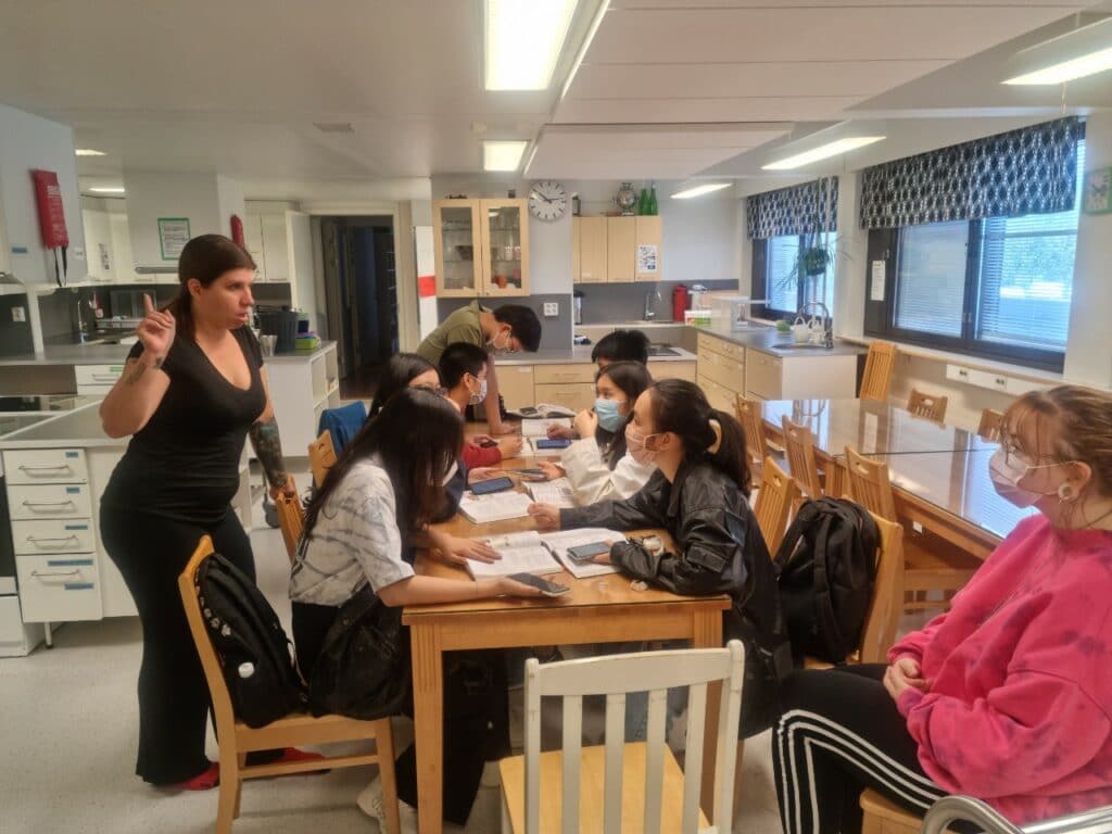 Study High School in Finland - Myths vs Facts 3 A group of students seated around a table in a classroom, some wearing masks. A teacher stands nearby, gesturing with one hand while addressing the students. Books and papers are spread out on the table. The classroom has large windows and a clock on the wall.