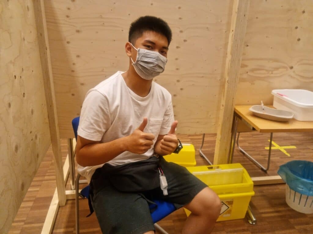 Study High School in Finland - Myths vs Facts 2 A person wearing a mask and casual clothing sits on a chair in a wooden booth. They are giving two thumbs up. Nearby, there are yellow bins and a table with trays.