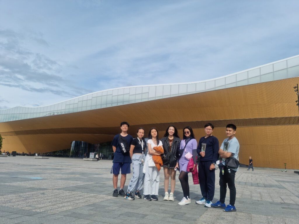 Celebrating Helsinki's Oodi Library - A Modern Marvel for Students and Culture Enthusiasts 6 A group of seven people stand in front of a modern architectural building with a wavy roof and wooden facade. The sky is partly cloudy, and they are posing on a large open plaza.