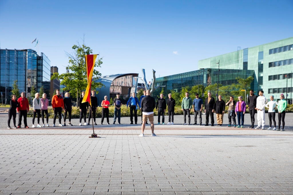 Finding my very own Finest Future in Finland 6 A group of people stand in a semi-circle on a paved outdoor area under a clear blue sky. A person in shorts and a jacket stands facing them, and a small flag is in the foreground. Modern buildings are visible in the background.