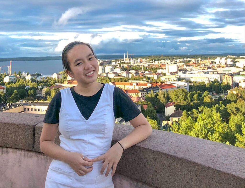 Finding my very own Finest Future in Finland 2 A person in a white shirt and dress stands smiling against a stone railing. Behind them is a scenic view of a city with trees, buildings, and a lake under a cloudy sky.