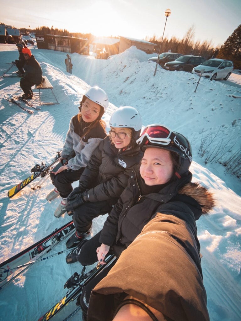 Finding my very own Finest Future in Finland 5 Three people in ski gear sit on a snowy slope. They are smiling and wearing helmets and goggles. Skis are attached to their boots. The sun is setting in the background, casting a warm glow over the scene. Other skiers and parked cars are visible nearby.