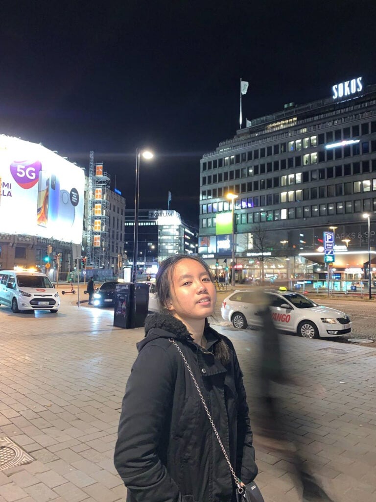 Finding my very own Finest Future in Finland 4 A person stands on a city street at night, with bright billboards and parked vehicles in the background. The scene is illuminated by streetlights, and buildings are visible in the distance.