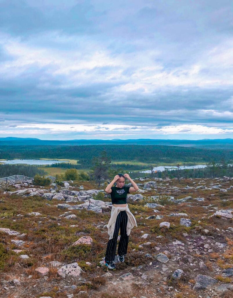 Finding my very own Finest Future in Finland 3 A person stands on a rocky hilltop surrounded by a vast landscape with green forest, winding rivers, and distant mountains under a cloudy sky. They are wearing casual outdoor clothing and appear to be adjusting their hair.